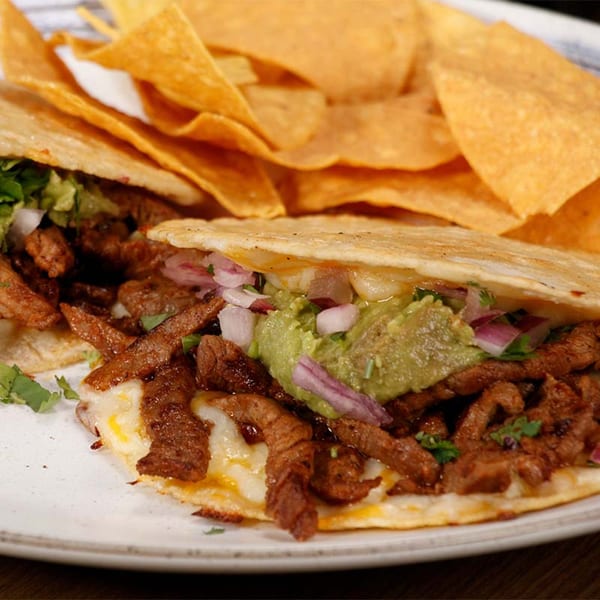 Two El Mexicano Tacos & Tequila tacos with steak, guacamole, onions, and cheese, served with tortilla chips.