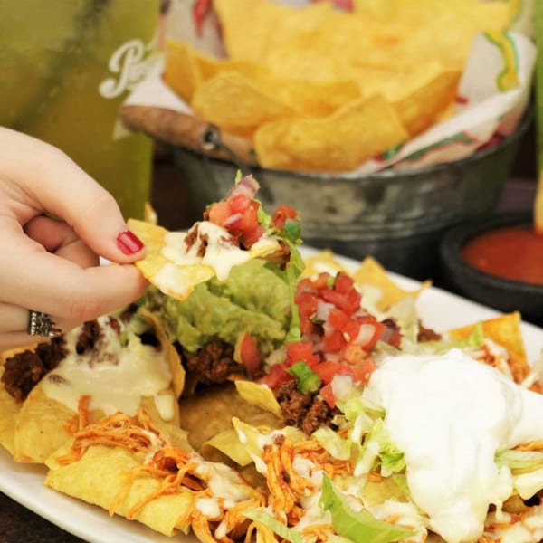 A hand with red nail polish picks up a chip from a large plate of El Mexicano nachos.