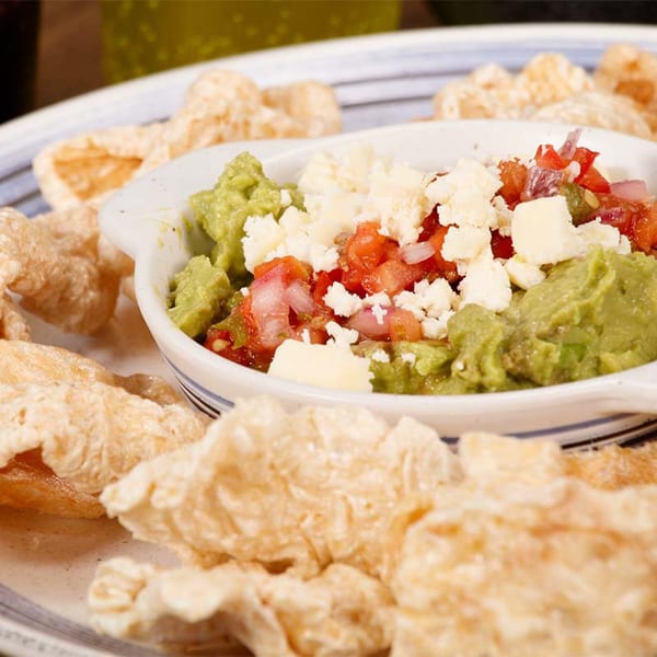 Guacamole and pico de gallo topped with cheese, surrounded by crispy chicharrones on a plate.